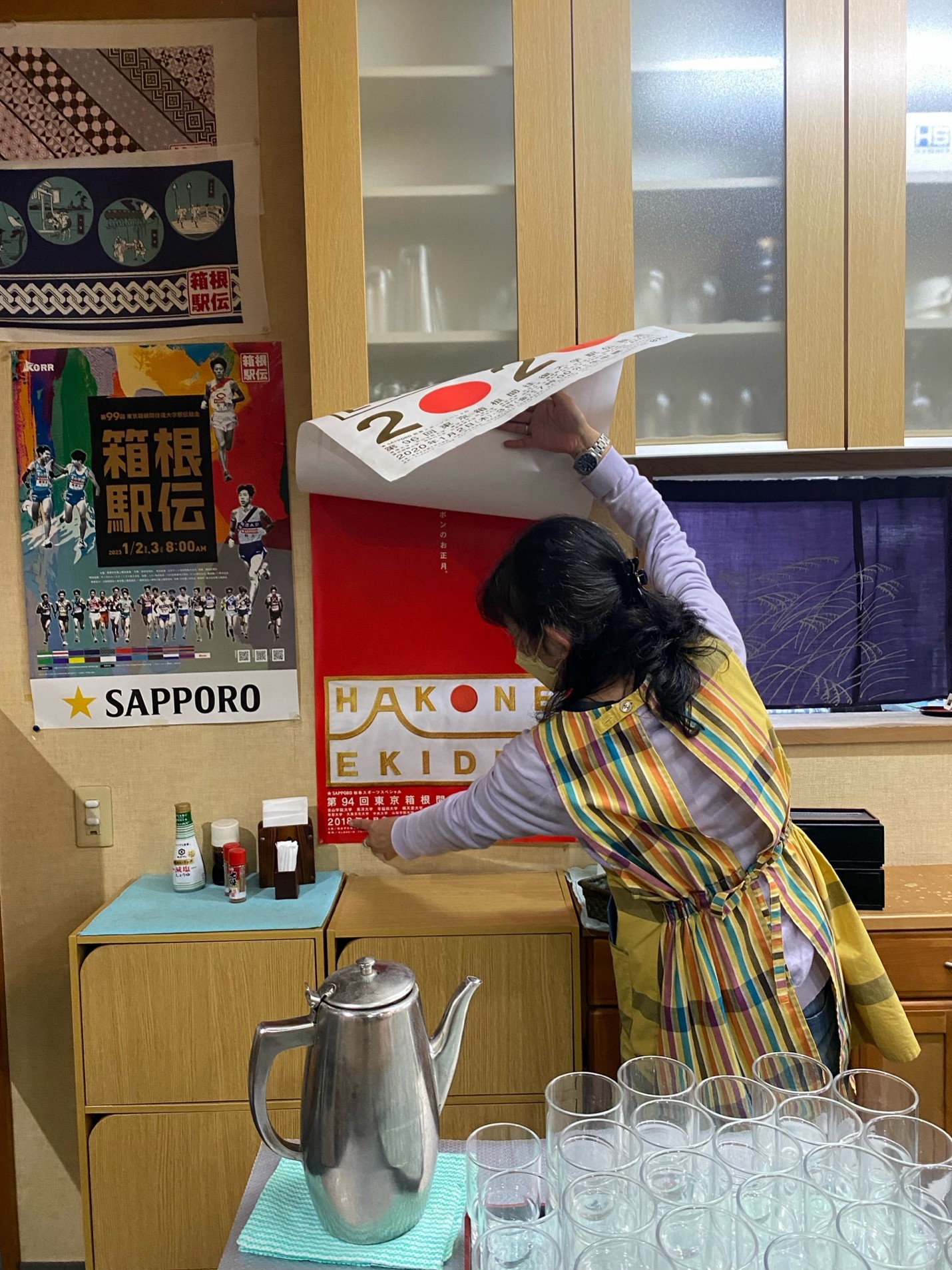 The portrait photograph depicts a black haired person (woman) from the back, wearing a yellow apron with blue, red and black stripes. At the lower quarter of the image in the front is a table with glasses and a coffee pot. In the background is a hanging kitchen cupboard on the right two thirds and on the left hang three posters. One is a colourful poster depicting people in movement like running. In the middle of that one poster is a black canvas with golden Japanese lettering. The person in the apron holds up a poster and points to another poster displayed on the wall.
