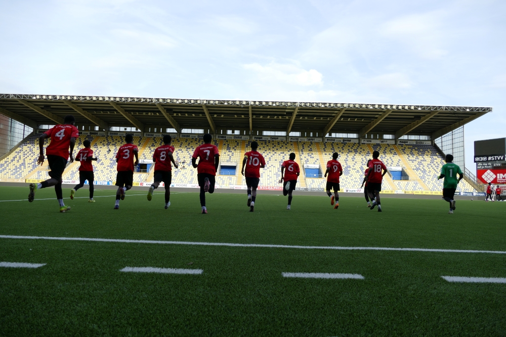 4, 14, 3, 8, 7, 10, 6, 9, 19 and 1 are the numbers displayed in white on the back of the red shirts of the people forming the horizontal centre of the photograph taken from the sidelines of a football field. In the background, one sees the sky and the yellow and blue stands that are flooded with sunlight. The players running across the field are in the shade.