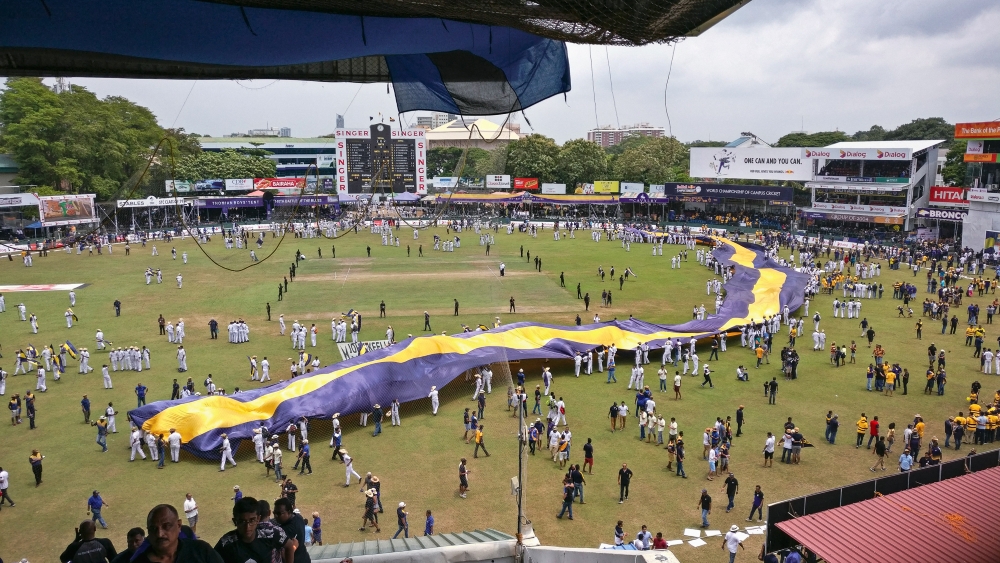 Image of a cricket pitch from the top of the viewer tribune. The field is full of people, but not crowded. From the back of the green, centre right of the photograph, people carry a very long banner that circles the main cricket pitch to the front left of the image. The green is also surrounded by stands and billboards of different heights.
