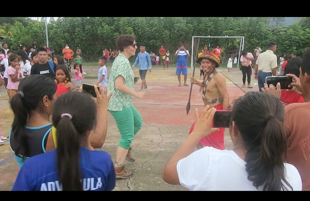 This is a scene on a concrete football pitch surrounded by tropical plants. On the pitch a group of people dance. The Indigenous performers dance barefoot and wear traditional red attire. One white looking person wears green trousers, a green and white shirt and hiking boots.