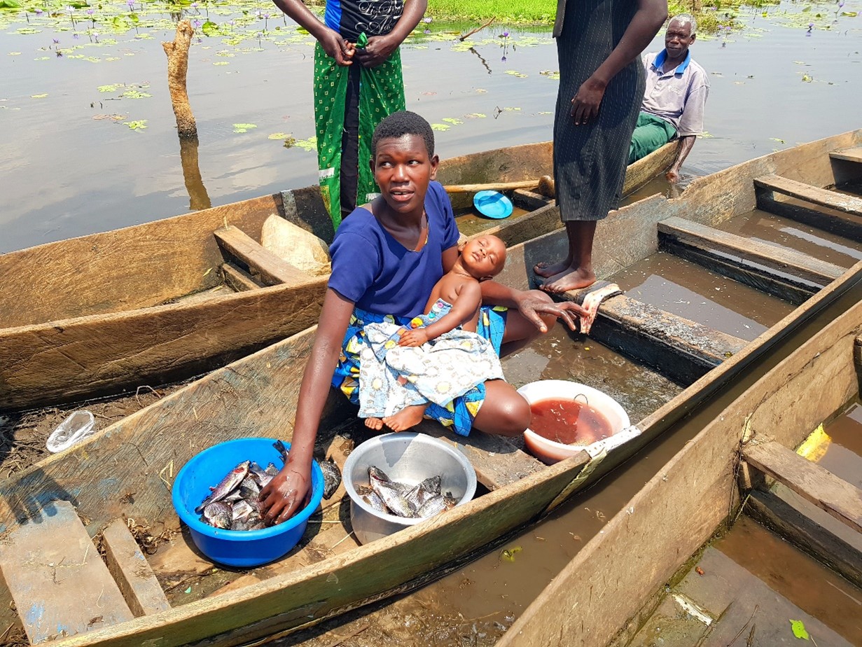 A woman cares for her baby while preparing fish for home consumption and sale on the River Nile in Apac district, Northern Uganda (2022).